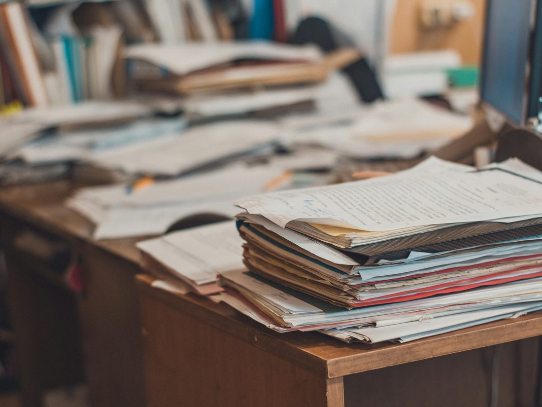 A wooden desk buried under stacks of paper, files and books.